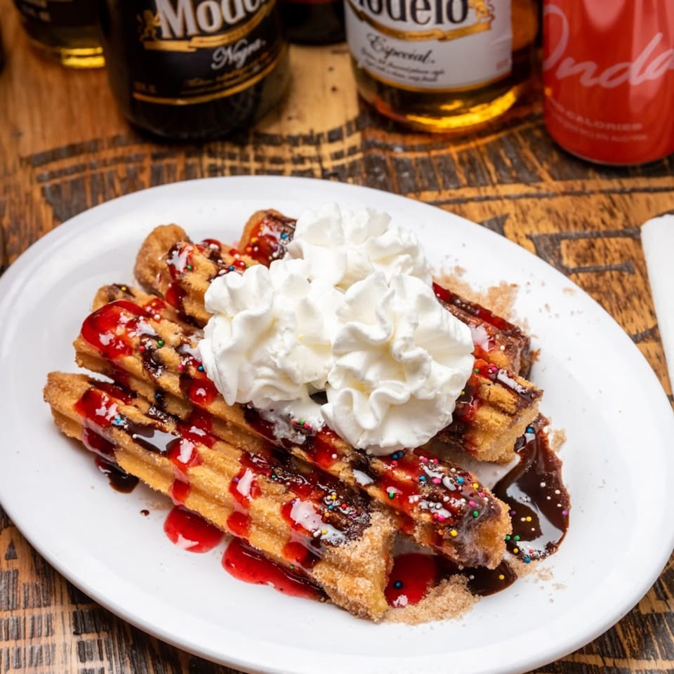 Churros with whipped cream, chocolate, strawberry sauce, and sprinkles on a white plate.